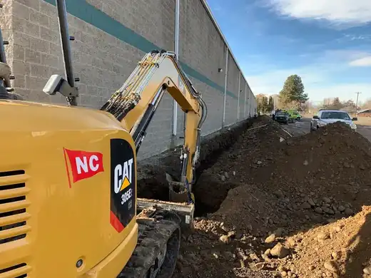 A mini-excavator trenching a service line along the side of a commercial building