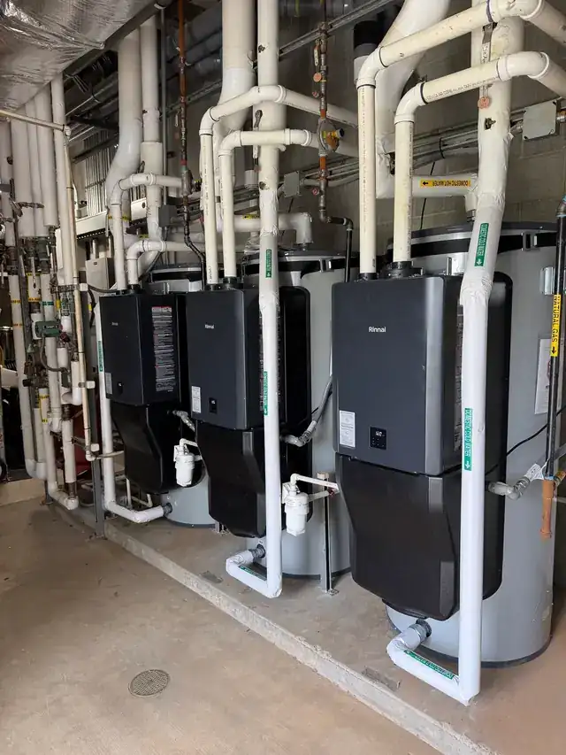 Three condensing tankless water heaters on a rack over storage tanks, with labelled lines and insulated piping