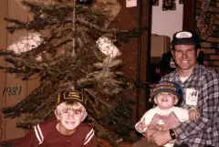The Burke family beside a Christmas tree marked 1981, a father in a cap with two young boys